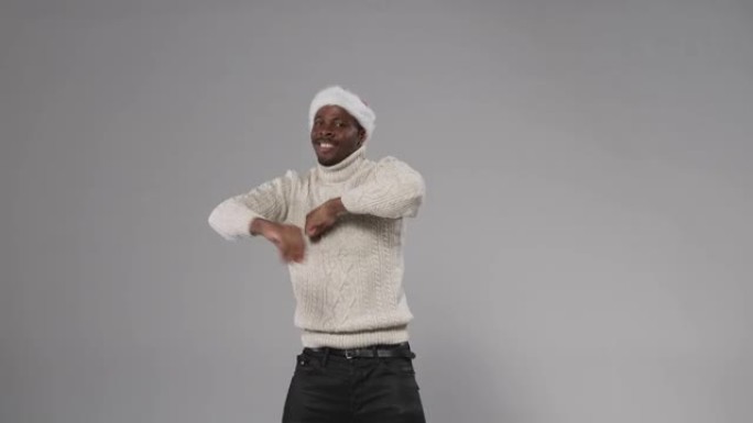 一个戴着圣诞帽和羊毛针织毛衣的黑皮肤男人在灰色背景上跳舞