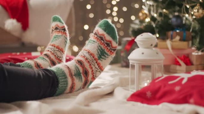 一个穿着圣诞节图案的袜子的男人坐在圣诞树附近的毯子上，移动双腿