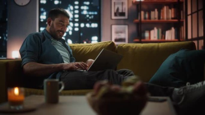 晚上，英俊的白人男子在黑暗舒适的客厅的沙发上休息时，在笔记本电脑上工作。自由职业者在家工作。浏览互联