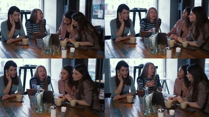 四个快乐兴奋的年轻美丽多样的女性朋友坐在现代咖啡店的餐桌上随意交谈。