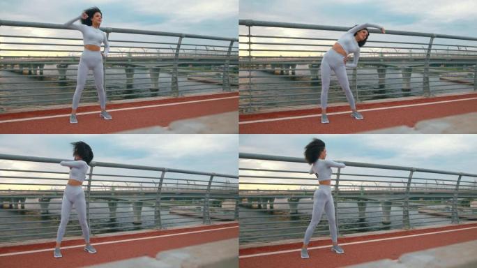 一位年轻健康的女跑步者穿着白色运动服，卷发，正在桥上热身。