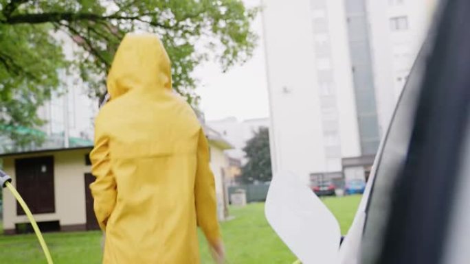 年轻的女性在雨中跳舞。开朗的年轻女子在雨中跳舞在正在充电的电动汽车旁边