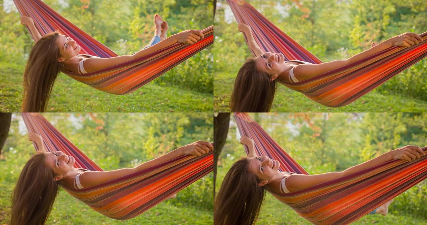 快乐的年轻女子在后院树下的吊床上摇摆，玩得开心，对着镜头微笑