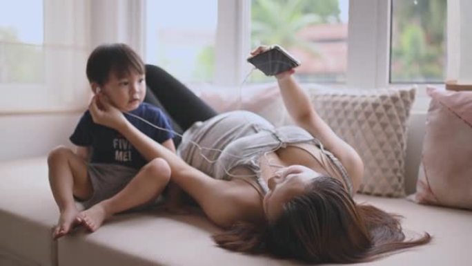 亚洲华裔孕妇躺在白天的床上放松，与儿子一起享受智能手机上的音乐