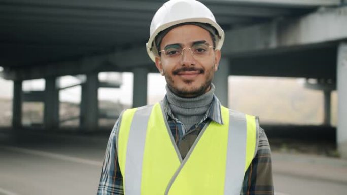 穿着建筑制服的快乐阿拉伯家伙站在城市建筑区微笑