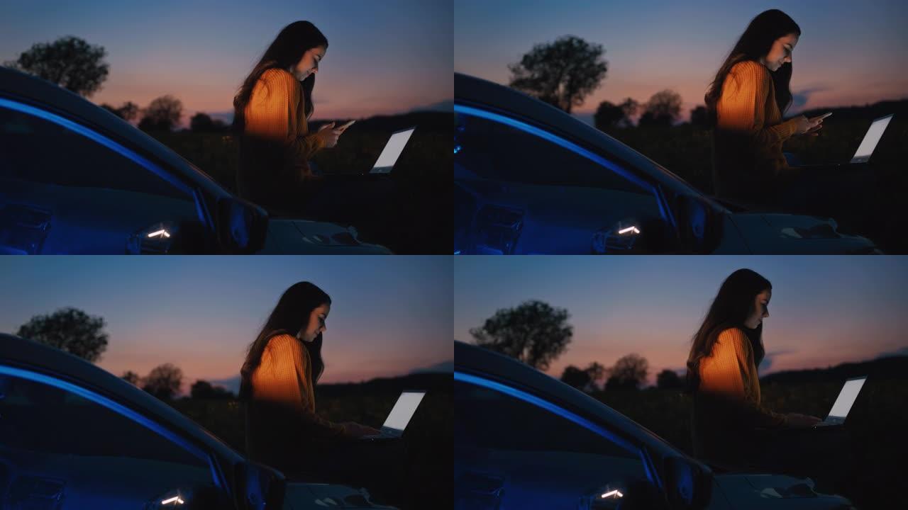 一个中年黑发女人坐在汽车引擎盖上时使用笔记本电脑和手机的慢动作，背景为黄昏天空