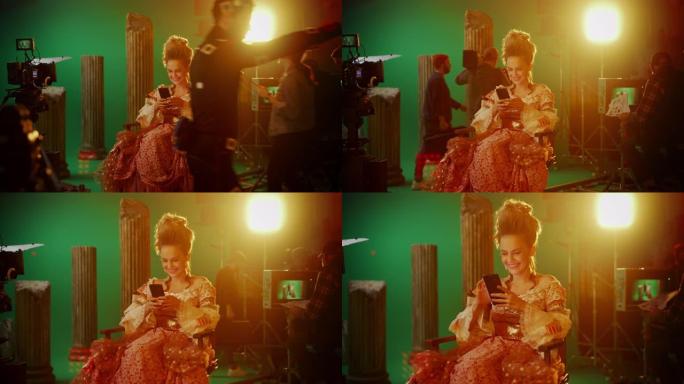 在古装戏剧电影中: 美丽的微笑女演员穿着文艺复兴时期的连衣裙，坐在椅子上，使用背景为绿屏的智能手机。