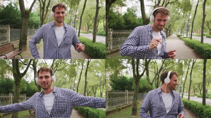 一个年轻无忧无虑的快乐男人的真实镜头是在阳光明媚的日子里戴着白色耳机听音乐和在绿色城市公园跳舞的乐趣