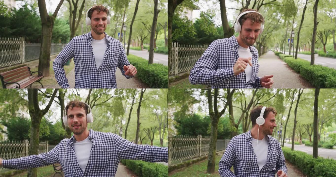 一个年轻无忧无虑的快乐男人的真实镜头是在阳光明媚的日子里戴着白色耳机听音乐和在绿色城市公园跳舞的乐趣