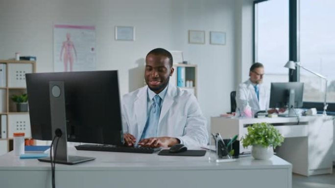 快乐微笑的非洲裔美国医生正在一家诊所的电脑上工作。穿着白大褂的医生正在医院办公室的桌子后面浏览病史。