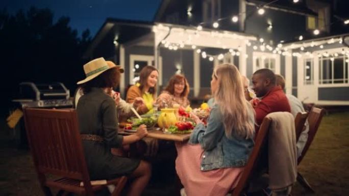 一群多民族的人玩得开心，互相分享故事，在户外晚宴上吃饭。缩小一个家庭在一个温暖的夏日夜晚聚集在他们家