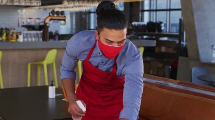 混合种族的男性咖啡馆工作人员戴着口罩消毒桌子