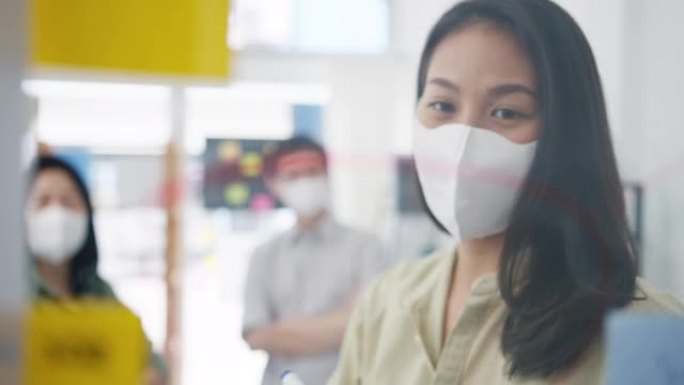 亚洲年轻商人讨论商业集思广益会议，在办公室工作时，他们一起分享数据并用医用口罩书写玻璃墙。