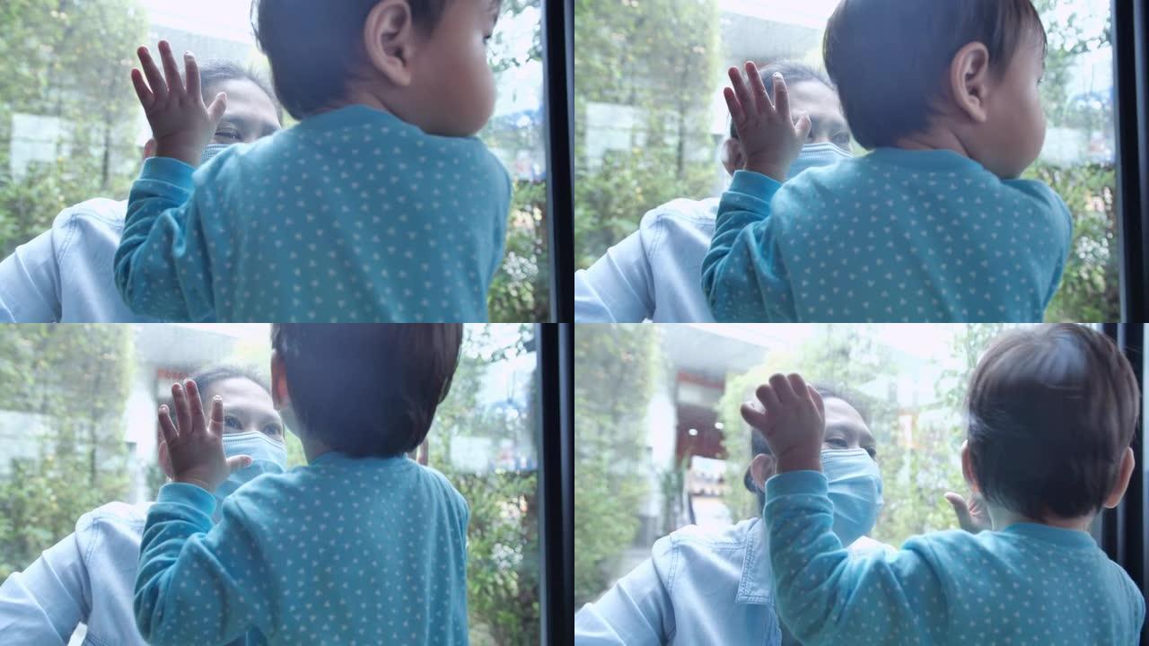 戴着防护口罩的年轻女性和12个月大的男婴通过玻璃窗触摸手掌，玻璃窗在社交距离期间将他们分开，以防止冠