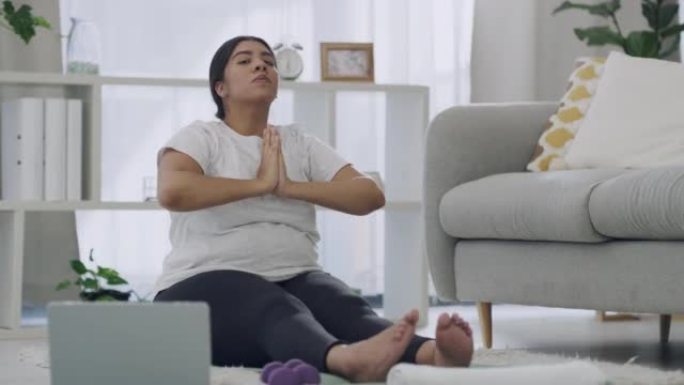 放松的女人在笔记本电脑上数字瑜伽课。健康的黑发女性在现代客厅地板地毯上冥想。年轻微笑的女性在她的休息