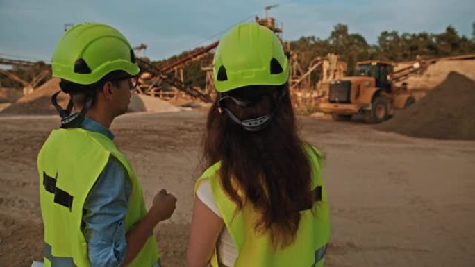 手持慢动作拍摄的两名采矿工程师指着一个装有挖掘机的采矿坑，戴着防护装备