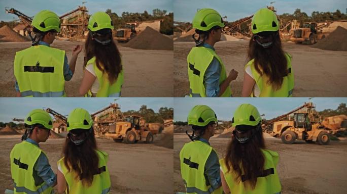 手持慢动作拍摄的两名采矿工程师指着一个装有挖掘机的采矿坑，戴着防护装备