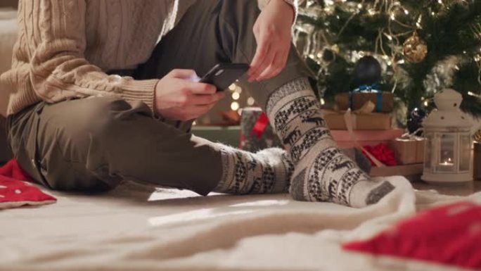 一位穿着圣诞袜的年轻女子坐在圣诞树附近的毯子上，在手机上输入一条消息。