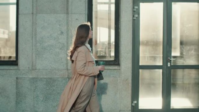 侧视图年轻漂亮的老板女商人穿着正式的米色外套，带着笔记本电脑沿着建筑墙壁慢动作行走。