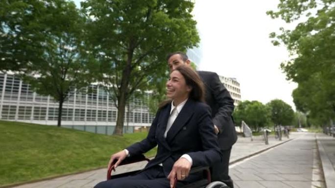 电影拍摄的快乐商人离开办公室后，在城市公园用轮椅推着他的女同事。企业、残疾人、友谊、团队合作、合作的