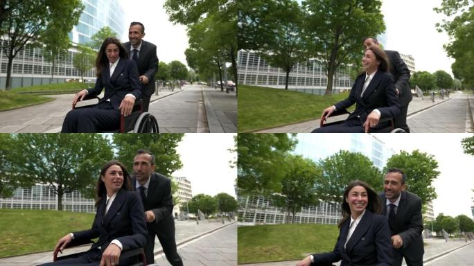 电影拍摄的快乐商人离开办公室后，在城市公园用轮椅推着他的女同事。企业、残疾人、友谊、团队合作、合作的