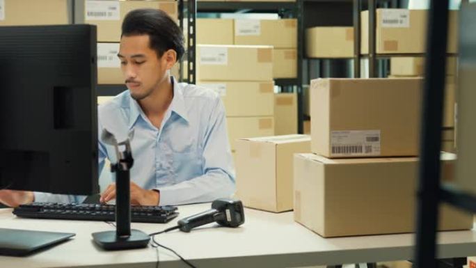 快乐的亚洲男人穿着正式的制服衬衫坐在办公桌前使用电脑和条码机扫描仪记录客户在线数据在纸箱仓库交货。创