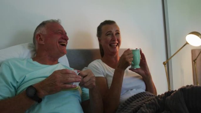 高加索高级夫妇在家里的床上喝咖啡杯时大笑