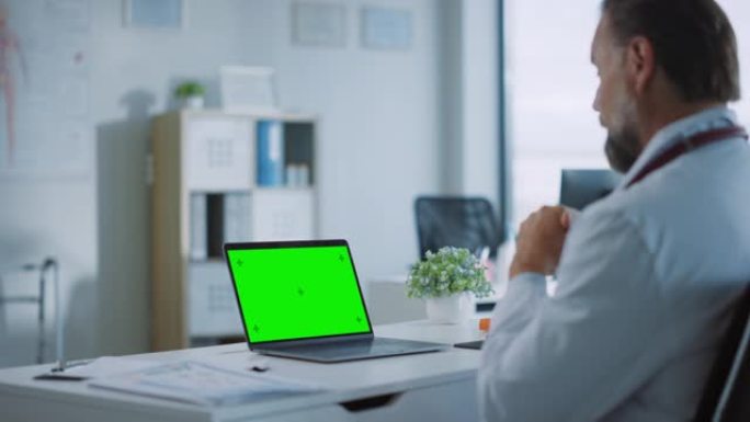 家庭医生正在健康诊所的绿屏显示的笔记本电脑上与患者进行视频通话。穿着实验室外套的助理正在医院办公室谈