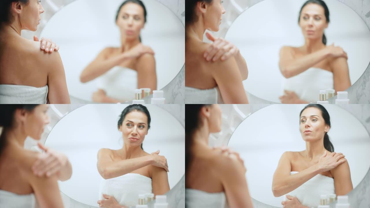 美丽的高加索女人在她完美的肩膀上涂上润肤霜，使皮肤柔软，照在浴室镜子里。快乐的女性享受她的美丽。健康