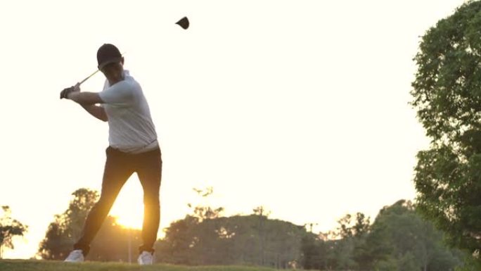 亚洲男子高尔夫球手挥杆高尔夫球杆，以慢动作击打日落背景的高尔夫球。