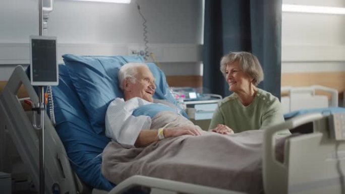 医院病房: 老人躺在床上休息，他有爱心的美丽妻子来访，支持他坐在旁边，手牵着手，幸福的夫妻交谈，微笑