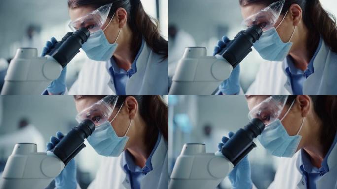 特写一位戴着口罩和护目镜的女科学家在显微镜下观察的镜头。用技术设备在现代实验室中研究分子样品的女性微