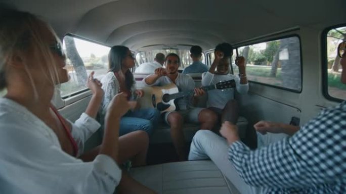 一群年轻的多民族朋友的慢动作在沿海公路上旅行时，在小型货车上弹吉他，唱歌和跳舞很有趣。