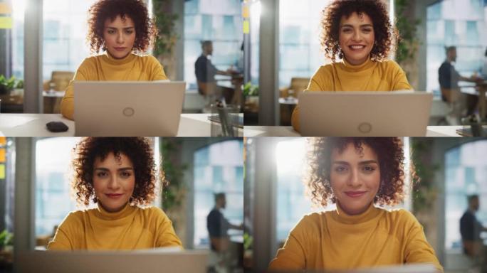 一位迷人的阿拉伯女性坐在创意机构的办公桌前的肖像。年轻时尚的经理，卷发微笑着，看着相机。在现代公司工