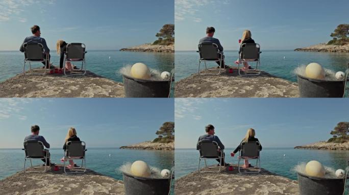夫妻，男人和女人坐在海边一块岩石上的野营椅上，杯子站在它们之间，女人在杯子里装满茶，两个人同时拿起杯