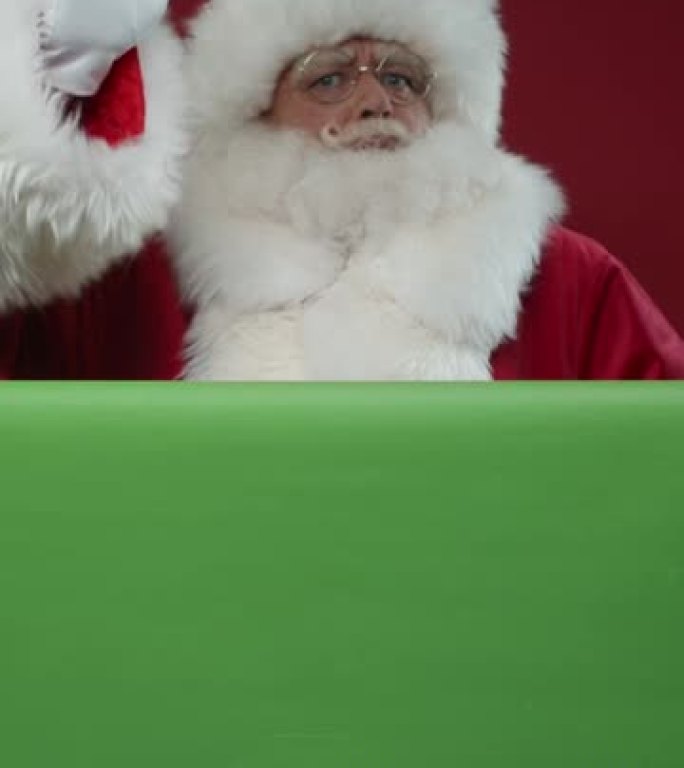 圣诞老人在绿色屏幕后面的红色背景下进入框架的垂直视频，对着镜头挥手，然后走开