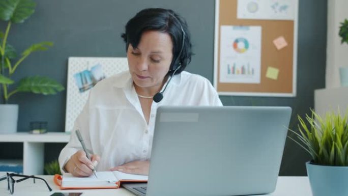 商务女士的慢动作戴着带麦克风的耳机书写并看着笔记本电脑屏幕