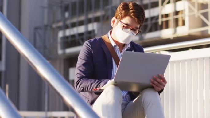 亚洲男子坐在公司公园的楼梯上戴着笔记本电脑戴口罩