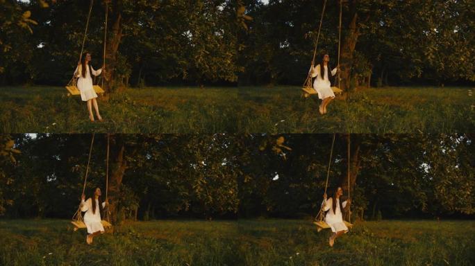 慢动作，手持镜头，一名穿着白色连衣裙的年轻成年女性摇摆，坐在悬挂在阳光充足的草地上的大树枝上的绳索秋