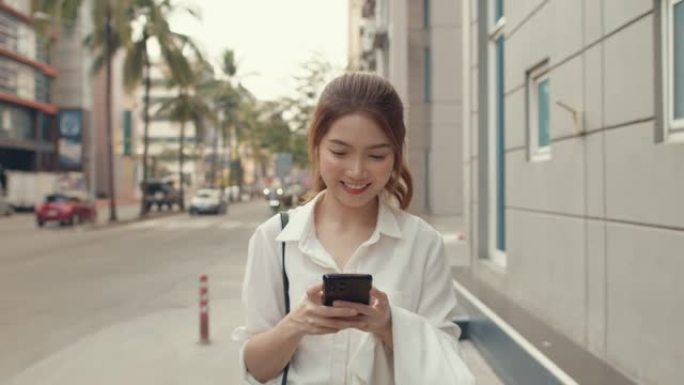 亚洲女商人穿着时尚办公服装，在城市现代城市中独自一人走在户外时，使用智能手机和打字短信。