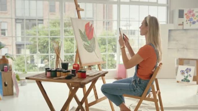 年轻的女画家在工作室用电话给一幅画拍照。艺术家为她在画布上画的花朵的最终产品拍照。女人为艺术品拍照