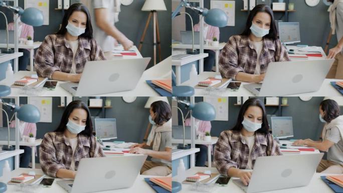 年轻的女商人戴着口罩，在办公室工作时使用笔记本电脑打字