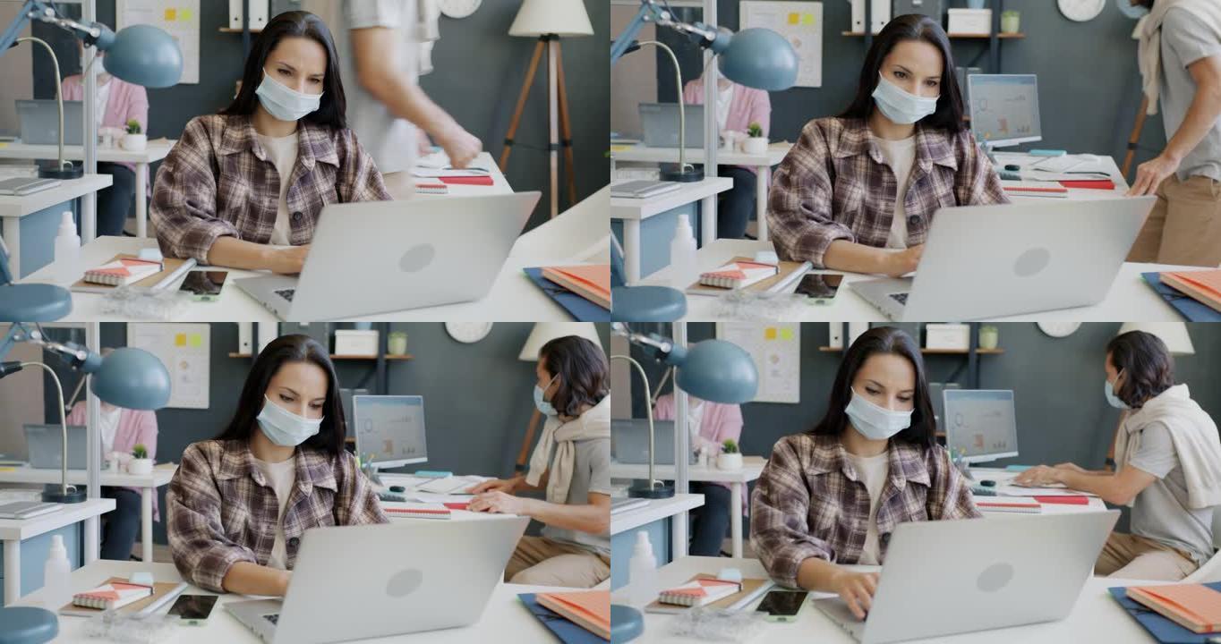 年轻的女商人戴着口罩，在办公室工作时使用笔记本电脑打字