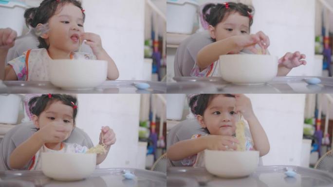 饥饿的亚洲蹒跚学步的孩子坐在椅子上，享受吃面条的乐趣，而与家人的女婴呆在家里，用手拿叉子在家里吃午饭
