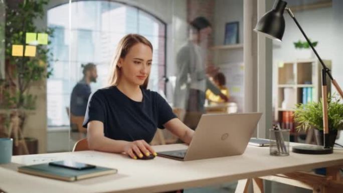 穿着黑色休闲装的美丽女性坐在创意办公室的办公桌前，在笔记本电脑上处理任务。年轻的创意专家为营销议程撰