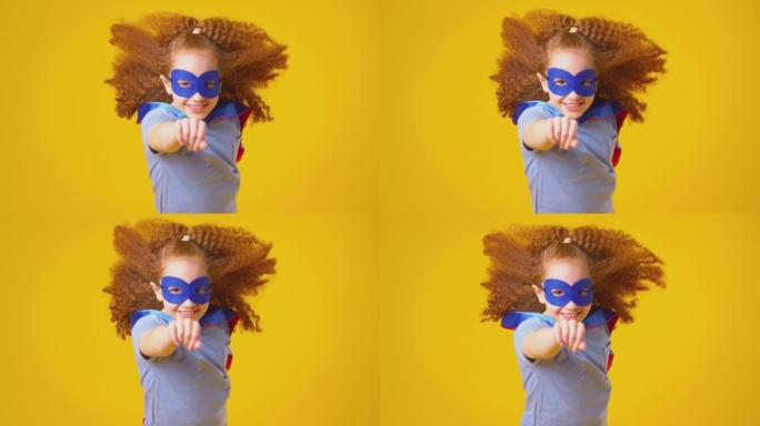 工作室拍摄的女孩打扮成漫画超级英雄戴着面具和斗篷假装在黄色背景下飞翔-慢动作拍摄