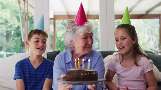 戴着党帽的高加索高级妇女在她的孙子坐在生日蛋糕上吹蜡烛