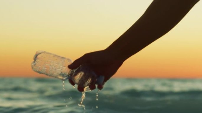 一只年轻志愿者的手的慢动作特写镜头正在从海里捡起一个塑料瓶，以保护日落时的环境。