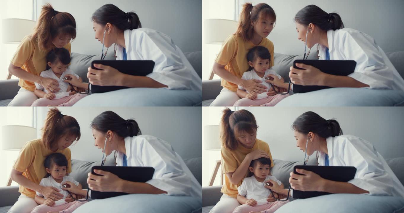 年轻的亚洲女儿科医生举行听诊器检查小女孩病人去看医生，母亲坐在家里客厅的沙发上。