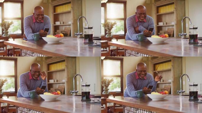 非裔美国人高级男子使用智能手机靠在厨房柜台上，摘下眼镜，微笑着
